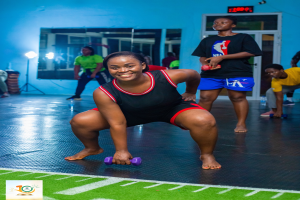 Eine Frau in einem Fitnessstudio führt eine Kniebeuge aus, während sie Hanteln hält und verschiedene Fitnessgeräte und Logos im Hintergrund zu sehen sind.