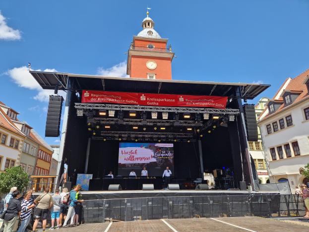 Gruppe von Menschen auf einer Bühne vor einem Uhrturm beim Kreissparkasse Gotha Musikfestival, mit Gebäuden, Bäumen und einem bewölkten Himmel im Hintergrund.