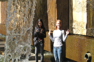 Zwei Frauen im Freien, eine posiert für ein Foto, während die andere eine Kamera hält, mit einem Wasserfall auf der linken Seite und einer Wand im Hintergrund.