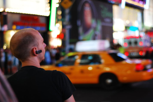 Eine Person in einem schwarzen T-Shirt und einem schwarzen Bluetooth-Headset steht vor einem gelben Auto auf einer Straße, mit anderen unscharfen Objekten im Hintergrund.