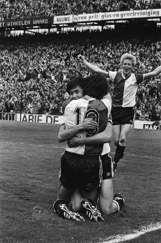 Drei Fussballspieler feiern ein Tor auf dem Boden, umarmen sich gegenseitig, mit einer jubelnden Menge und schwenkenden Fahnen im Hintergrund, dargestellt in schwarz-weiß.