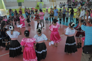 Eine Gruppe von Kindern in Kostümen tanzt in der Mitte des Bildes, während das Publikum von der rechten oberen Seite zusieht.