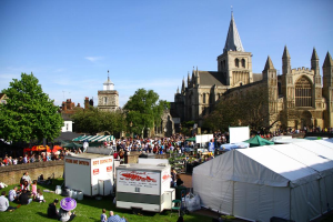 Historische Gebäude mit Bäumen davor, zusätzliche Gebäude und ein Turm im Hintergrund, ein Weg mit Menschen und Geschäften, und Personen, die auf dem Rasen in der Nähe der Geschäfte sitzen.