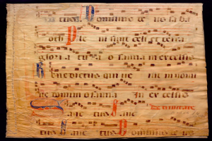 Ein illuminiertes Manuskript mit Text und Musiksymbolen auf einem Blatt Papier vor dunklem Hintergrund.