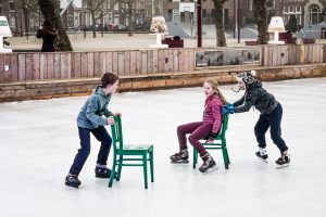 Kinder beim Skifahren im Vordergrund eines Spielplatzes, mit drei Kindern und zwei Stühlen in der Mitte und Gebäuden, Bäumen, Bänken, Pfählen und einem Basketballfeld im Hintergrund.