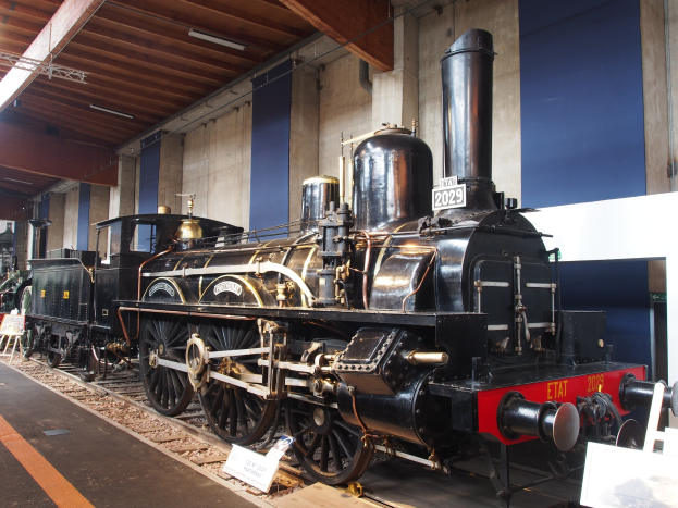 Alte Dampflokomotive in einem Museums Setting umgeben von Eisenbahnschienen, Informationsschildern und anderen Ausstellungen, mit einer Wand und beleuchteter Decke im Hintergrund.
