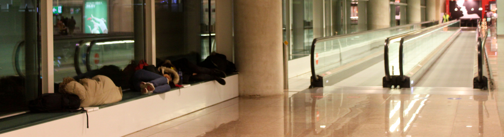 Menschen auf Bänken in einem Flughafen mit zwei geraden Rolltreppen im Hintergrund.