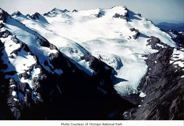 Majestätischer Berg mit einem großen Gletscher, umgeben von schneebedeckten Gipfeln und einem klaren blauen Himmel, mit Text unten.
