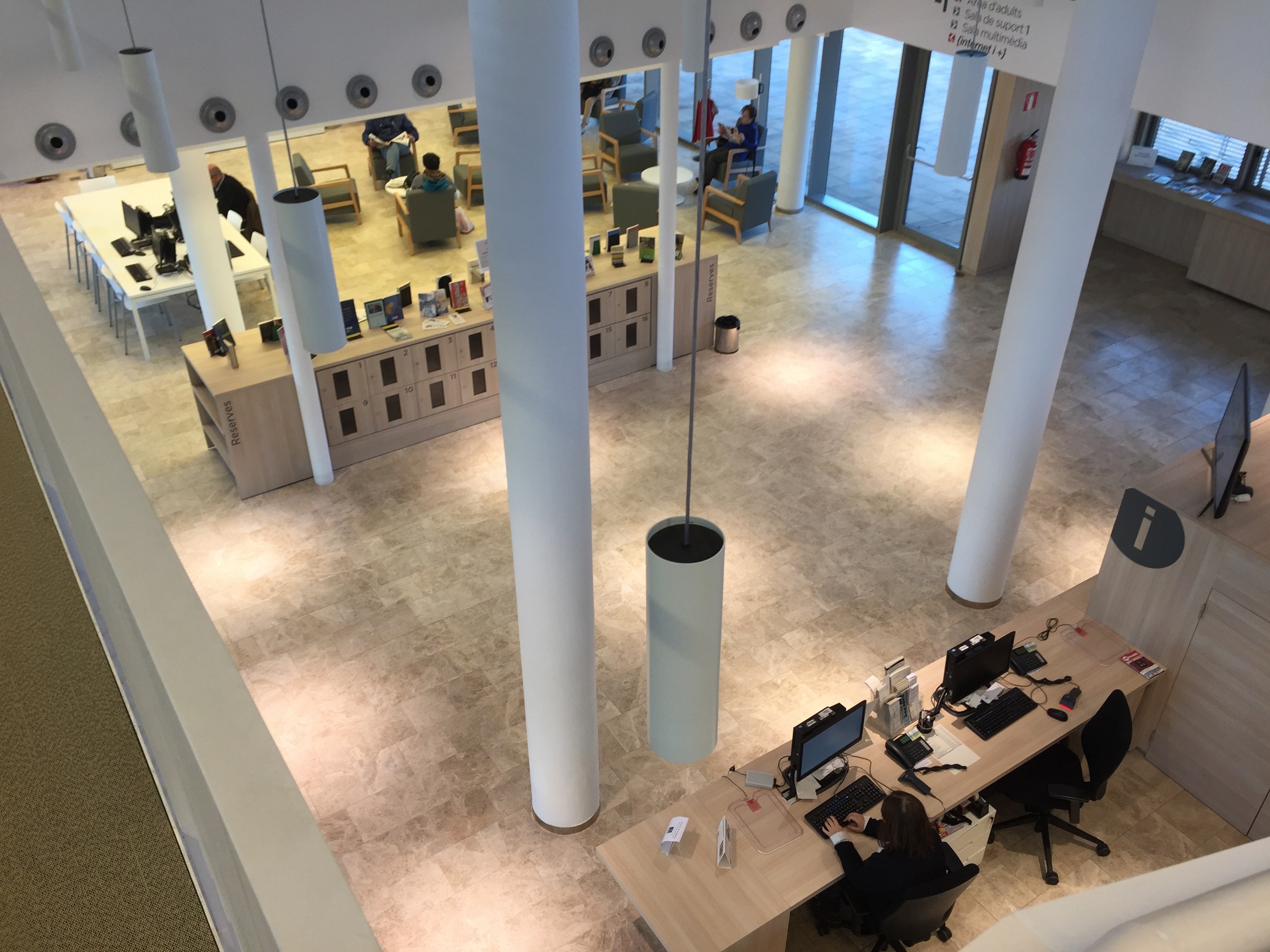 Großraumbüro mit Menschen an Schreibtischen mit Computern, Monitoren, Tastaturen und Papieren, mit Deckenleuchten und Säulen, einem Feuerlöscher rechts und einer Tafel mit Text.