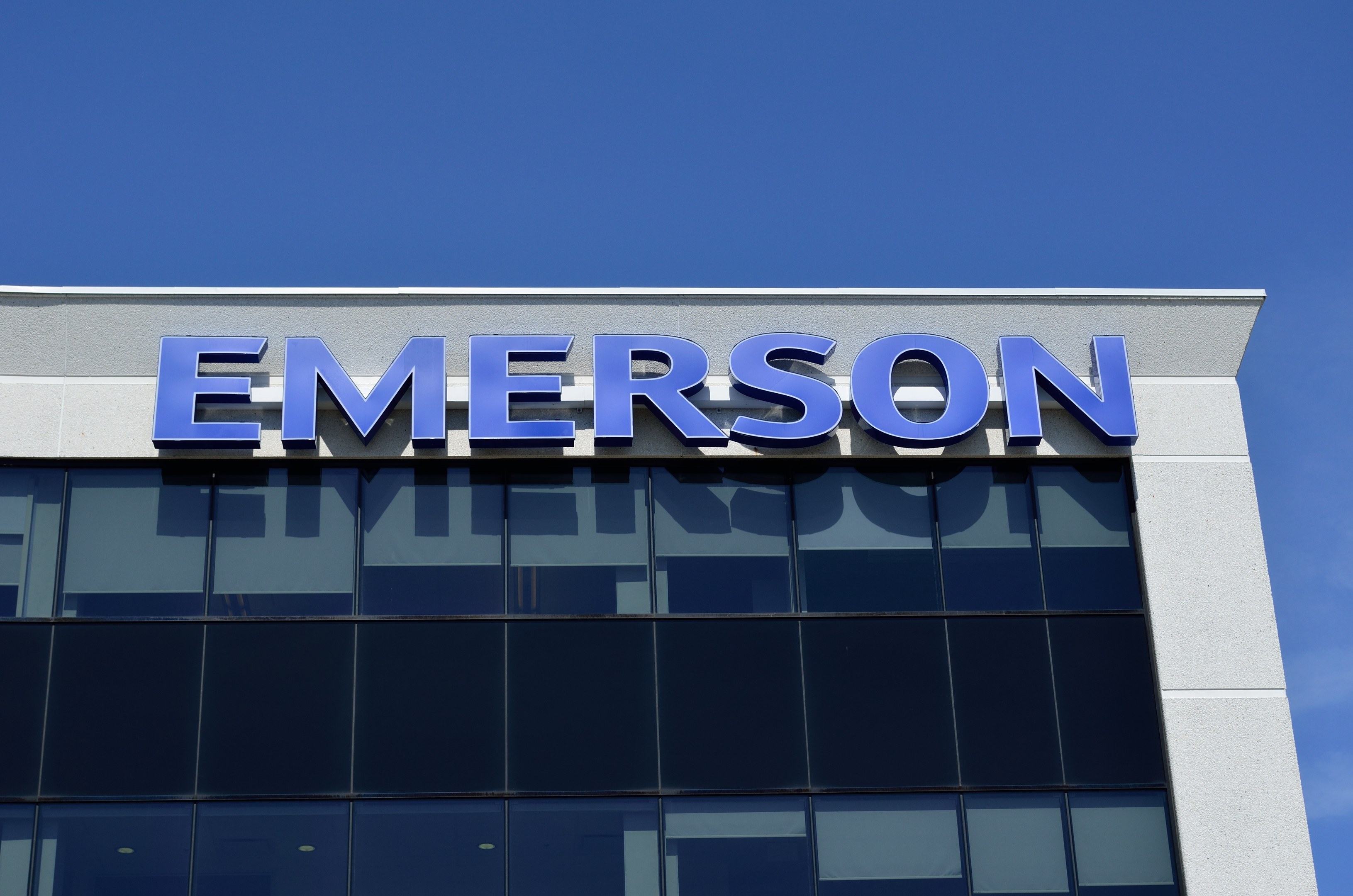 Emerson-Logo an einer Gebäudefassade mit dem Text "Emerson investiert 1,5 Milliarden USD in den USA" vor einem klaren blauen Himmel.