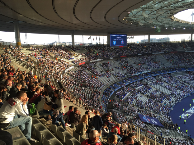 Große Menschenmenge in einem Stadion bei einem Fußballspiel mit einer Bühne, Fahnen und einem Bildschirm im Hintergrund, identifiziert als Allianz Arena in München, Deutschland.