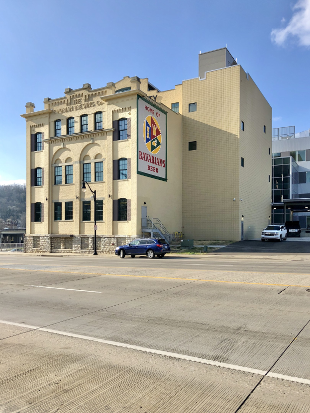 Großes industrielles Gebäude mit "Home of Bavarian Brewing Co." -Schriftzug, Straßenlaternen, Fahrzeugen, Bäumen, Hügeln und bewölktem Himmel im Hintergrund.