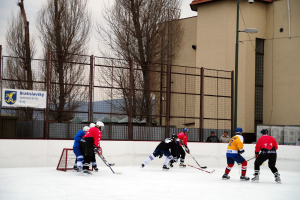 Menschen beim Eishockeyspielen auf einem Eis mit Gebäuden, Bäumen, einer Straßenlaterne, einem Schild und Zäunen im Hintergrund unter einem Himmel.