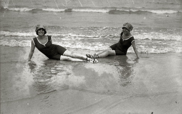 Zwei Frauen in Bikinis und Hüten, die in Schwarz-Weiß-Fotografie strahlend im Meer sitzen.