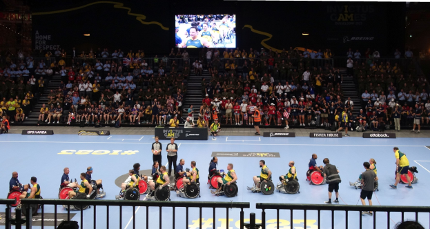 Eine Gruppe von Menschen in Rollstühlen auf einem Feld bei den Invictus Games Düsseldorf 2020, mit einer großen Zuschauermenge im Hintergrund und einer Treppe, die zu einem Bildschirm führt.