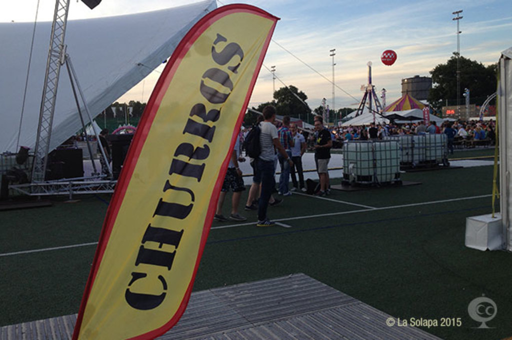 Ein buntes Faschingszelt mit Zelten, einem zentralen Mast, Menschen, B├Ąumen, Strommasten, Straßenbahnen auf der Stra├če und einem eisernen Stand auf der linken Seite.
