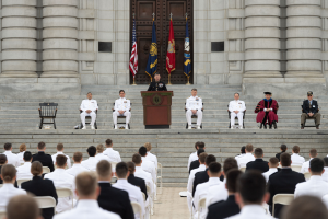 Gruppe von Menschen, die auf Stühlen vor einem Gebäude mit Säulen und einer Tür sitzen, mit einem Rednerpult und einem Mikrofon in der Mitte, Flaggen im Hintergrund und einem leeren Stuhl links daneben.