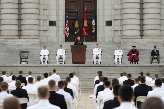 Gruppe von Menschen, die auf Stühlen vor einem Gebäude mit Säulen und einer Tür sitzen, mit einem Rednerpult und einem Mikrofon in der Mitte, Flaggen im Hintergrund und einem leeren Stuhl links daneben.