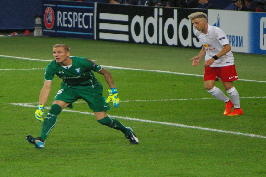Zwei M├Ąnner spielen Fussball auf einem saftig gr├╝nen Feld, umgeben von einer Menschenmenge, einige halten Kameras, mit Texttafeln im Hintergrund.