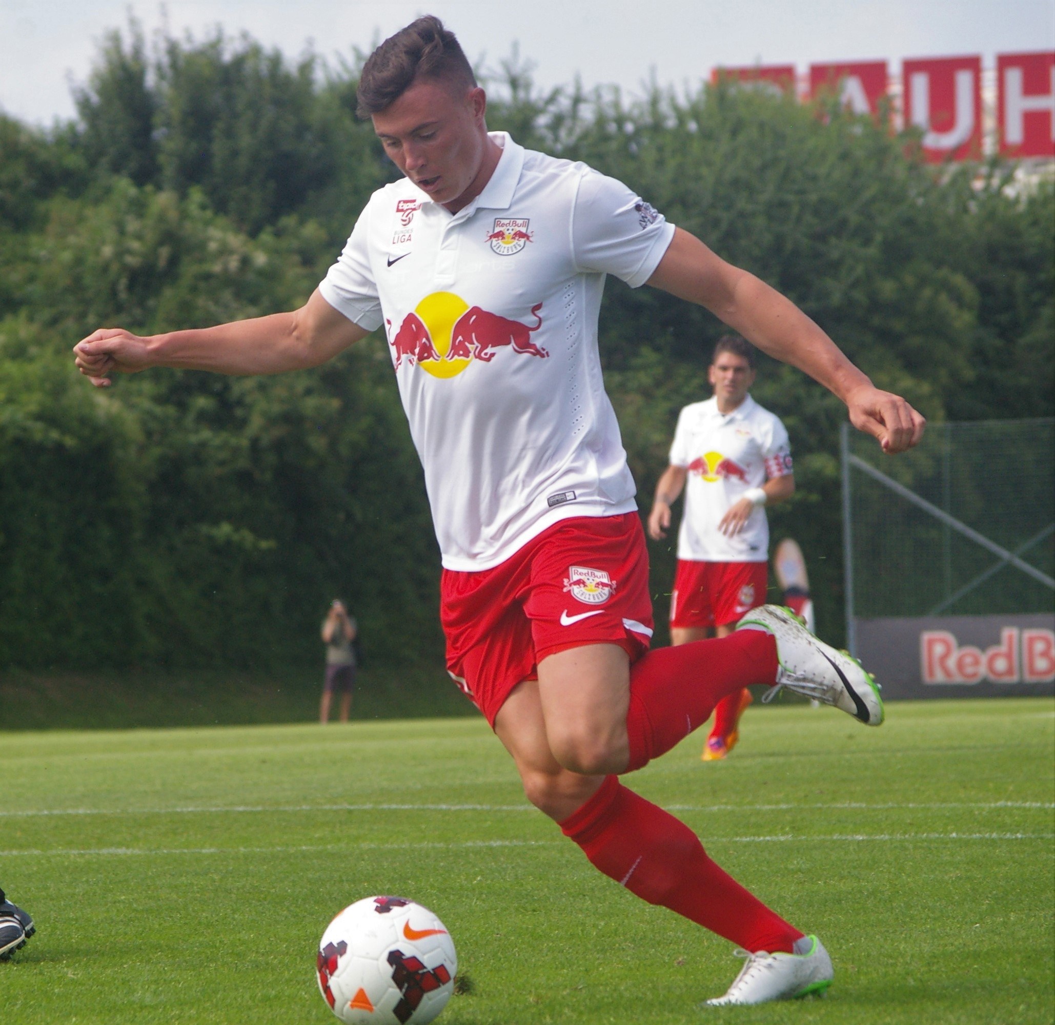 Ein professioneller Fussballspieler schießt einen Ball auf einem grünen Feld mit Bäumen und einem klaren blauen Himmel im Hintergrund, in der Nähe einer Tafel mit der Aufschrift "RB Leipzig."