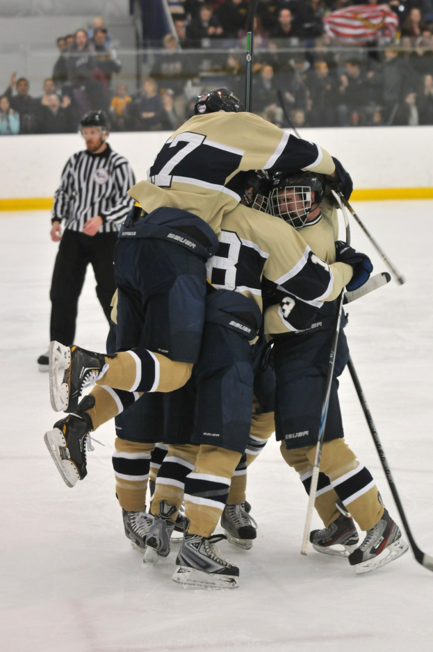Eine Gruppe von Hockey-Spielern in voller Ausrüstung drückt sich auf dem Eis zusammen, mit Zuschauern und einer Wand im Hintergrund.