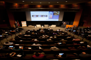 Ein mit sitzenden Menschen gefüllter Hörsaal, beleuchtet von zahlreichen Lichtern, mit Fahnen links, einem Podium rechts und einer Werbetafel links.
