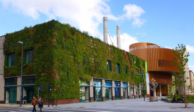 Ein großes Gebäude mit grüner Wand umgeben von Straßeninfrastruktur, Fahrzeugen, Fußgängern, Bäumen und einem bewölkten Himmel, Teil eines nachhaltigen Green Building-Projekts.