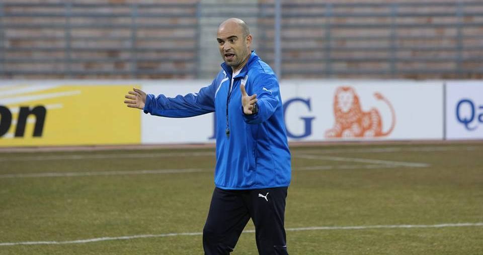 Ein Mann in einer blauen Jacke steht auf einem Fußballfeld während eines Trainings, mit entschlossener Miene und Tafeln mit Text und Logos im Hintergrund.