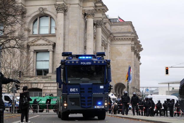 Gruppe von Polizisten vor einem großen Gebäude mit architectural details, Fahrzeuge auf der Straße, eine Person mit einer Kamera auf der linken Seite und Hintergrund-Elemente einschließlich Bäume, Verkehrszeichen, Flaggen und einen klaren blauen Himmel.