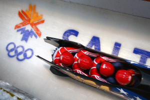 Ein Rodler in einem Schlitten bei den Olympischen Spielen, mit einer dekorierten Wand und Text im Hintergrund.