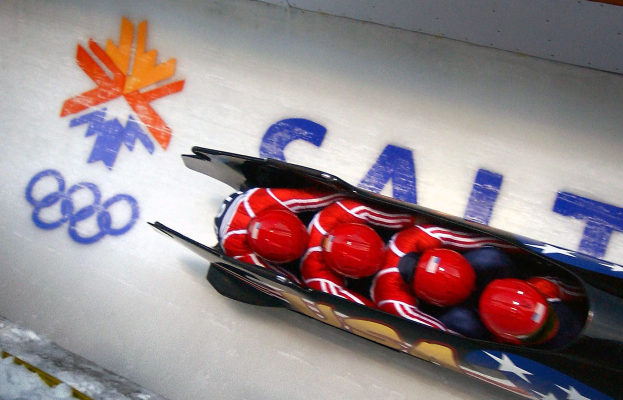 Ein Rodler in einem Schlitten bei den Olympischen Spielen, mit einer dekorierten Wand und Text im Hintergrund.