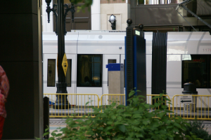 Frau auf der linken Seite des Bildes vor einem Zug auf einer Stadtstraße umgeben von Gebäuden, Straßenmöbeln und Grünflächen.