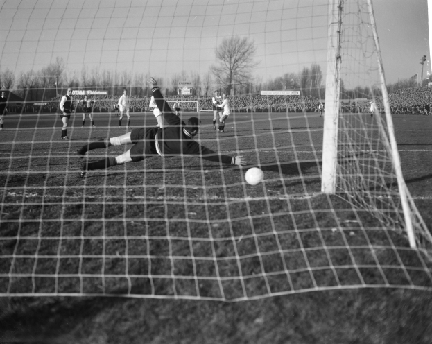 Schwarzes und weißes Foto eines Fußballtorwarts, der den Ball vor einem Tor abwehrt, umgeben von Spielern auf dem Boden und Hintergrundelementen wie Bäumen, Pfosten, Werbetafeln und einem klaren Himmel.