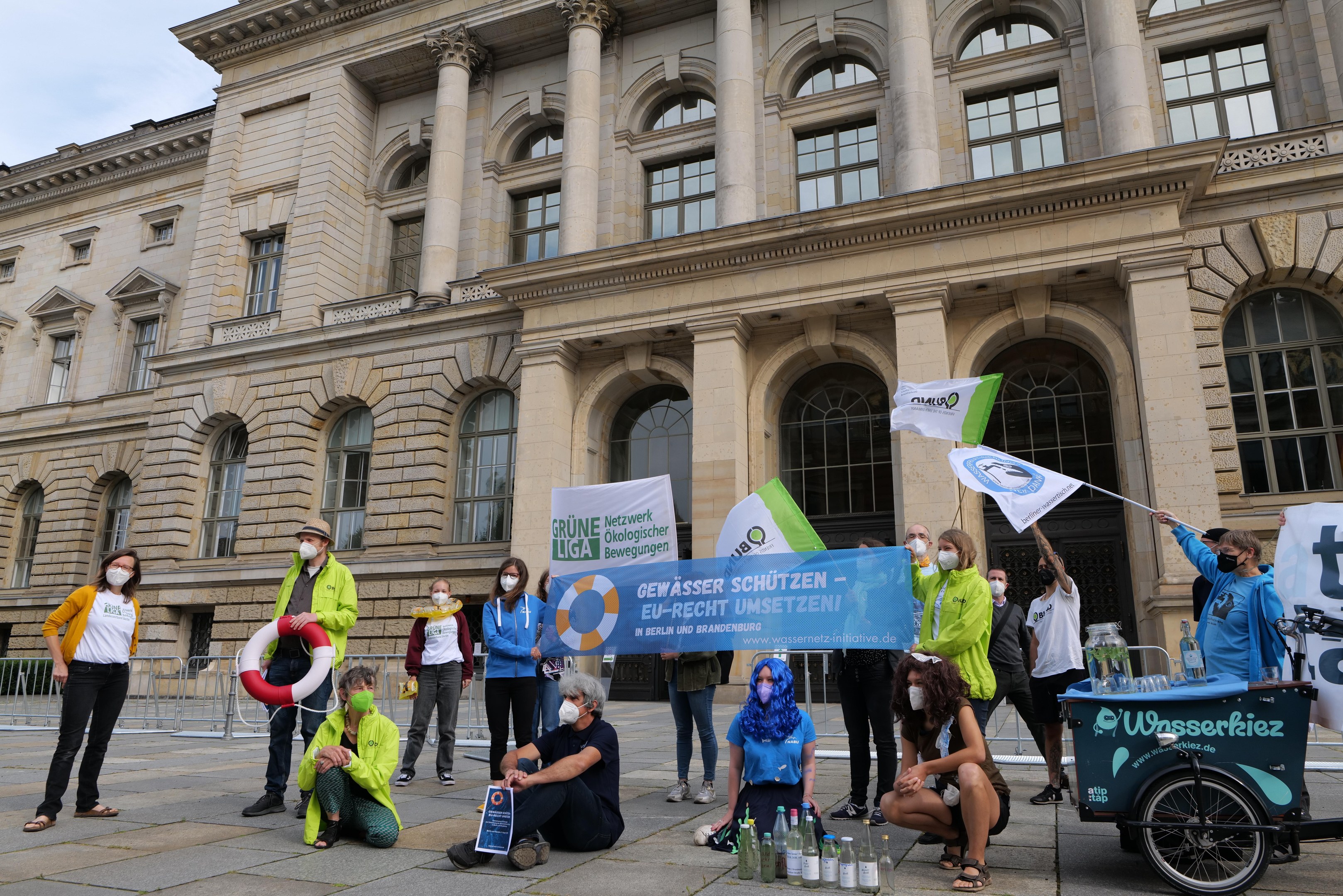 Eine Gruppe von Menschen in Masken mit Schildern vor einem Gebäude mit Säulen und Bögen während eines Klimastreiks in Berlin, mit einem Fahrzeug und einem Geländer im Hintergrund.