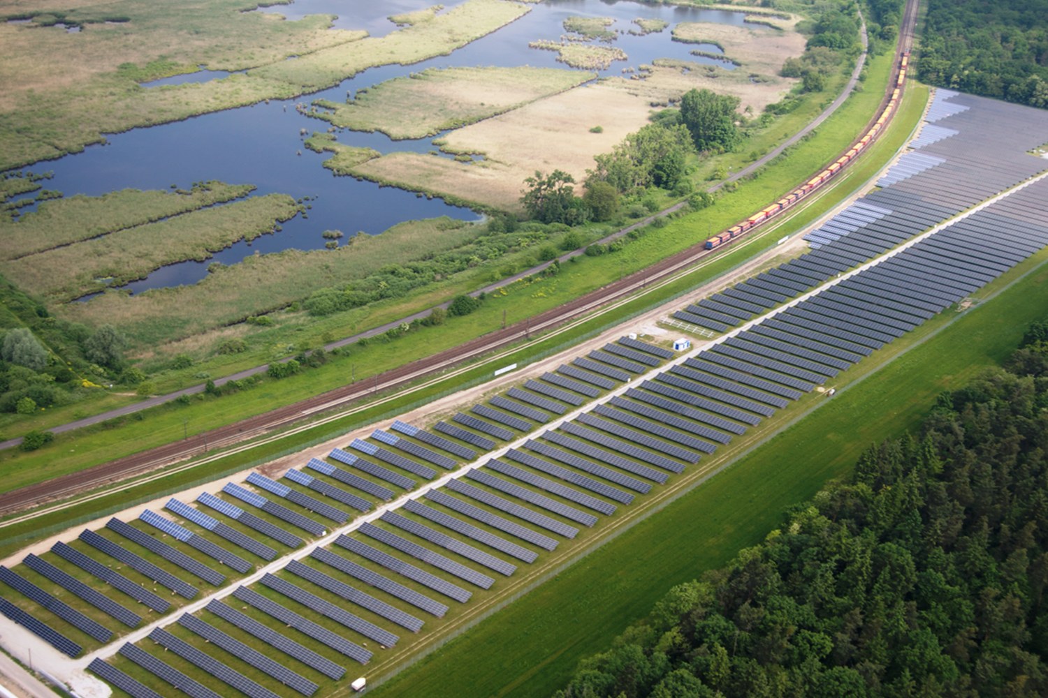 Luftaufnahme eines Solarparks mit Solarpanelen in einem Feld, umgeben von Bäumen, Gras, Pflanzen und Wasser, mit einem Zug auf einem nähergelegenen Bahngeleis.