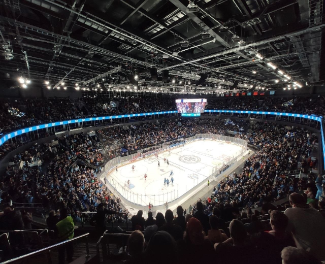 Eishockeyspiel in einer großen Arena mit Zuschauern auf den Rängen und Personal am Boden, mit Geländern, Deckenbeleuchtung und einem Bildschirm, identifiziert als O2 Arena in London.