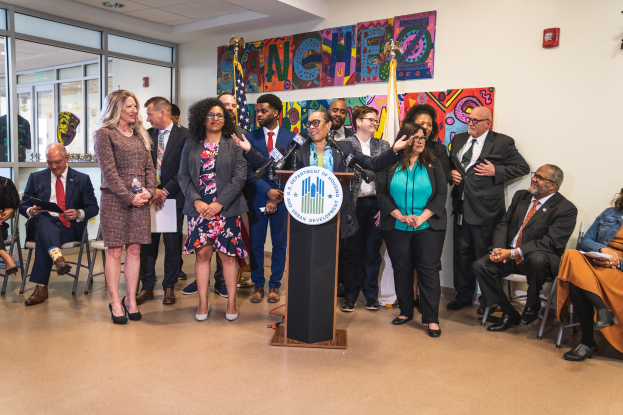 Gruppe von Menschen in einem Raum mit einem Podium, einem Mikrofon und einer Tafel mit der Aufschrift "Sancho-Stadtrat tritt dem Stadtrat bei", umgeben von Stühlen, Fahnen, einem Feuerlöscher und Deckenleuchten.