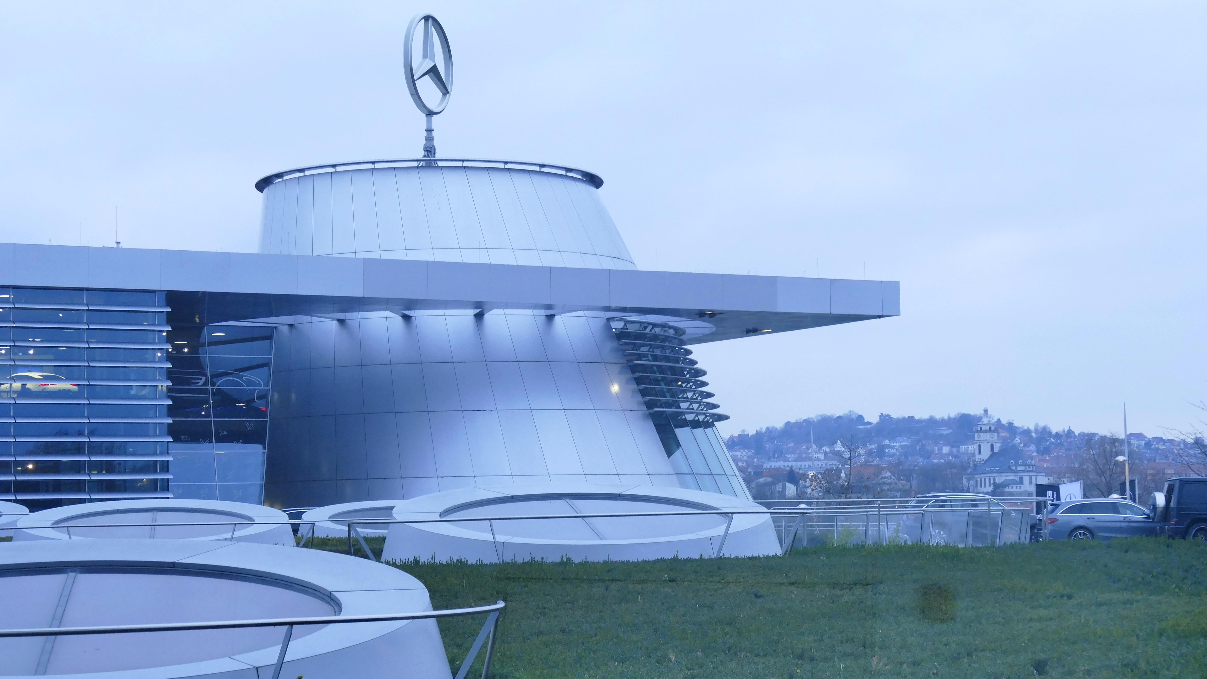 Das Mercedes-Benz Museum in Frankfurt, Deutschland, zeigt sein Logo auf dem Dach, umgeben von Gras und Geländern, mit Fahrzeugen vorne und Bäumen im Hintergrund unter einem klaren blauen Himmel.