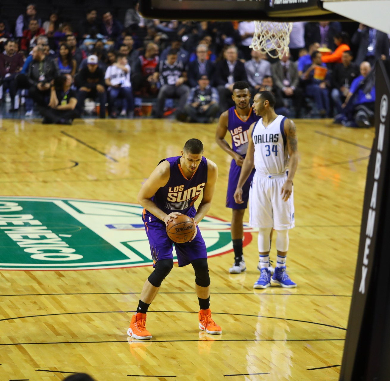 Basketball-Spiel im Gange zwischen den Phoenix Suns und den Dallas Mavericks, mit Spielern auf dem Feld, einer den Ball haltend, Anzeigetafel über dem Korb sichtbar und Zuschauern in den Rängen.