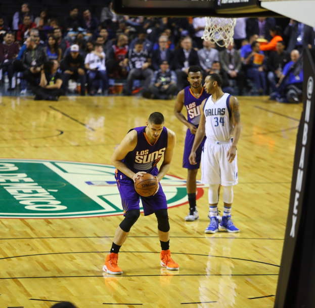 Basketball-Spiel im Gange zwischen den Phoenix Suns und den Dallas Mavericks, mit Spielern auf dem Feld, einer den Ball haltend, Anzeigetafel über dem Korb sichtbar und Zuschauern in den Rängen.