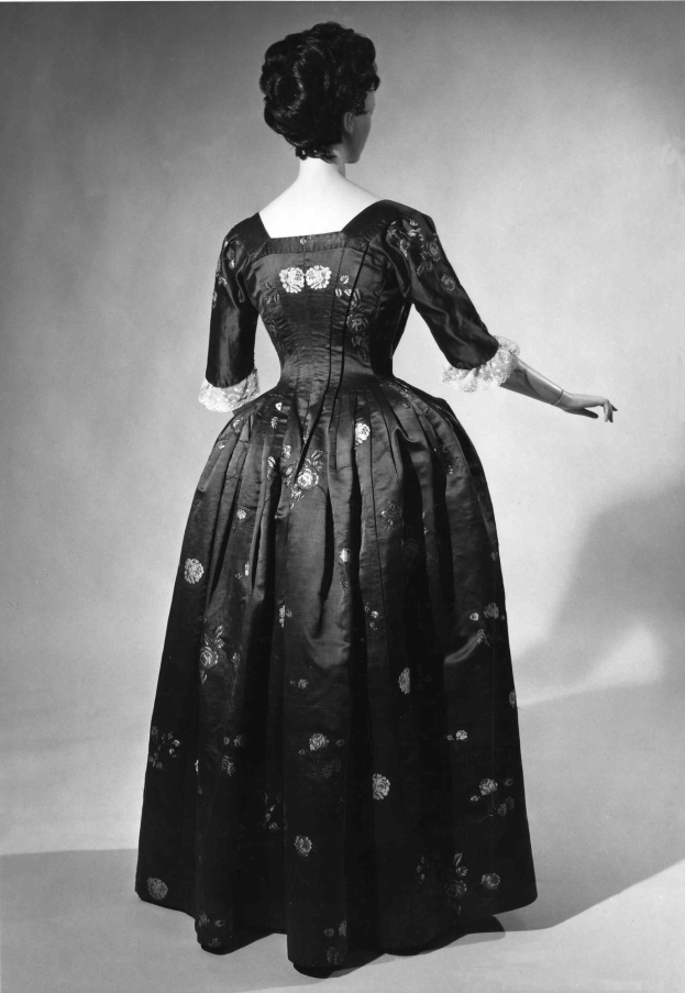 Eine Frau in einem 1950er-Langkleid mit engem Oberteil und vollem Rock steht mit gefalteten Händen, hochgestecktes Haar, schwarze Pumps und dezentem Make-up.