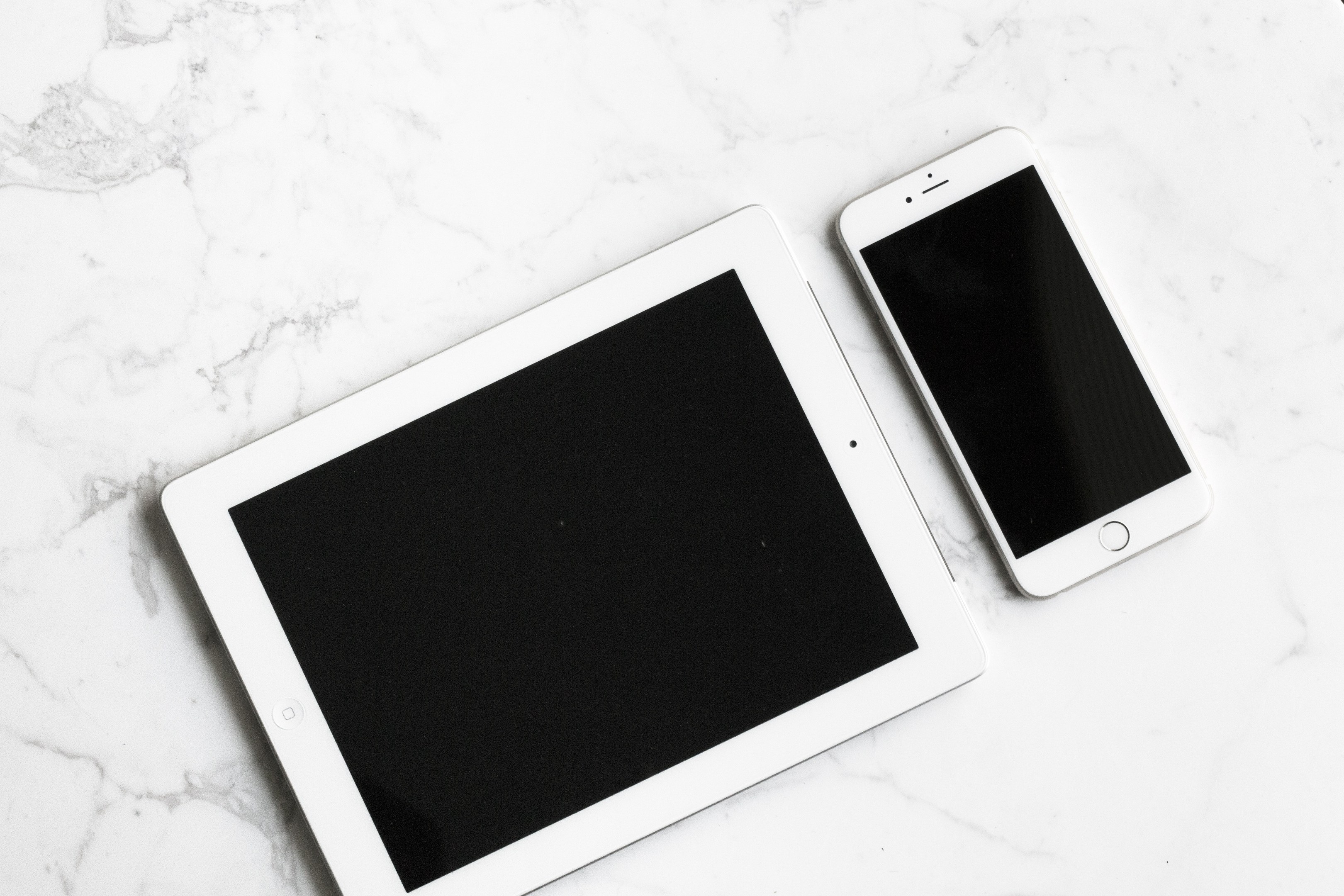 Zwei Tablets, ein iPad und ein iPhone, nebeneinander auf einer weiße Marmorfläche plaziert.