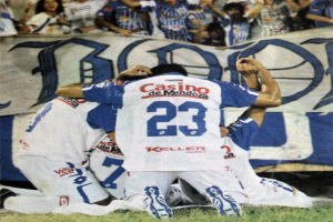 Fußballspieler feiern ein Tor auf dem Feld mit jubelnden Zuschauern und einer Fahne im Hintergrund.