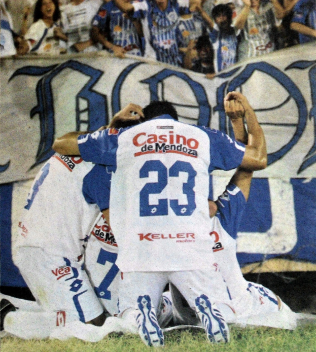 Fußballspieler feiern ein Tor auf dem Feld mit jubelnden Zuschauern und einer Fahne im Hintergrund.