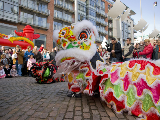 Ein lebendiges chinesisches Neujahrsfest in Amsterdam mit einem Löwen tanzen im Vordergrund und einer Menge Menschen drumherum, Gebäude und Laternenpfähle im Hintergrund und einem klaren blauen Himmel.