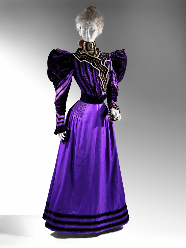 Eine Schaufensterpuppe trägt ein bodenlanges lila Abendkleid mit einem schwarzen und goldenen gemusterten Rock, der mit goldener Stickerei und Perlen verziert ist, eine taillierte Taille mit Spitzenbesatz und eine gestylte Frisur mit zarten Ohrringen.