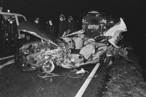 Schwarzes und weißes Foto eines auf der Straße liegenden Wagens mit mehreren Menschen, einige mit Helmen, drumherum und einem Fahrzeug im Hintergrund.
