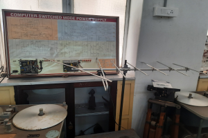 Ein unordentlicher Elektronik-Workshop mit einer beschrifteten Leiterplatte auf einem Tisch, Metallstangen, einer wandmontierten Schalttafel, einem Fenster und Gegenständen in Schränken, was auf eine computergeschaltete Modus-Stromversorgung hinweist.