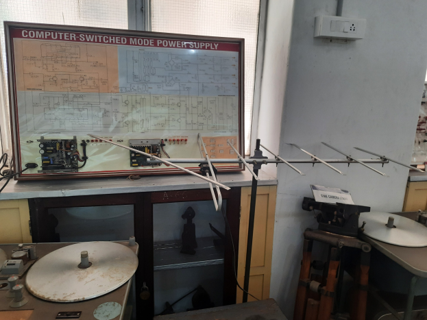 Ein unordentlicher Elektronik-Workshop mit einer beschrifteten Leiterplatte auf einem Tisch, Metallstangen, einer wandmontierten Schalttafel, einem Fenster und Gegenständen in Schränken, was auf eine computergeschaltete Modus-Stromversorgung hinweist.