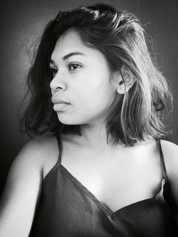 Eine selbstbewusste Frau in einem schwarzen Kleid posiert in einem Schwarz-Weiß-Foto, ihr glatter Bob und das natürliche Make-up unterstreichen ihre selbstbewusste Haltung an einer Wand.